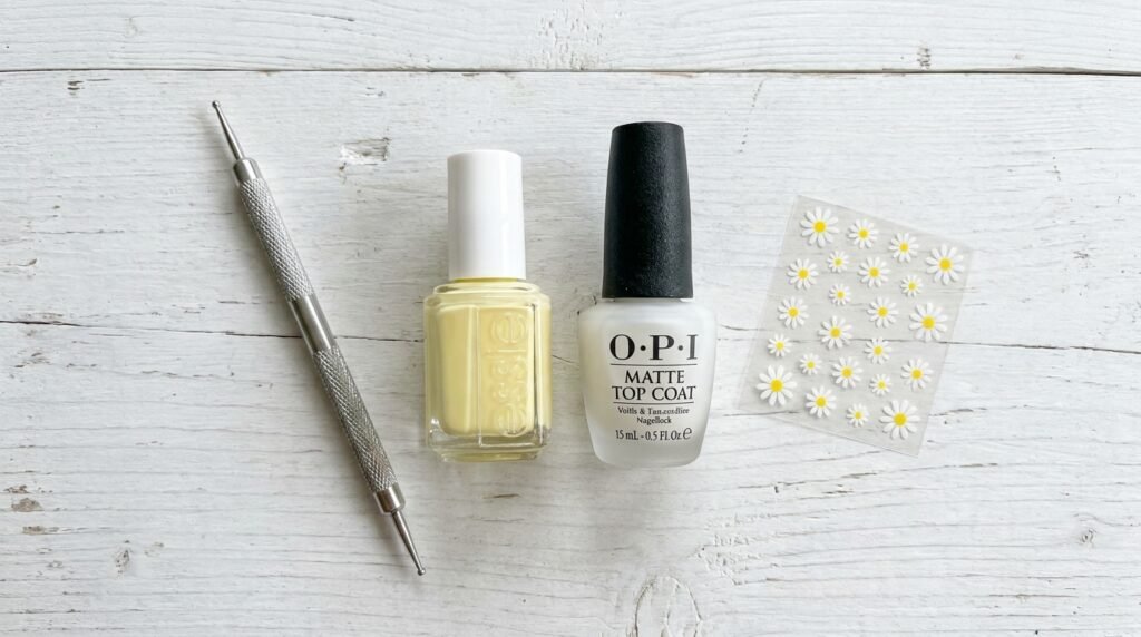 Overhead view of DIY nail art tools including pastel yellow polish, a metal dotting tool, and white flower decals.