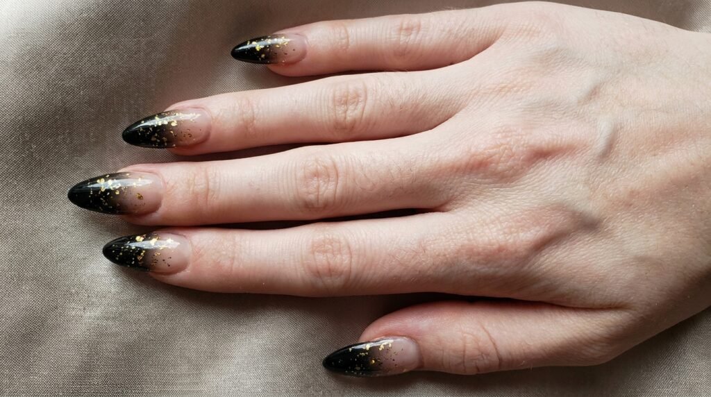Almond nails with milky nude bases fading into black tips with gold glitter accents.