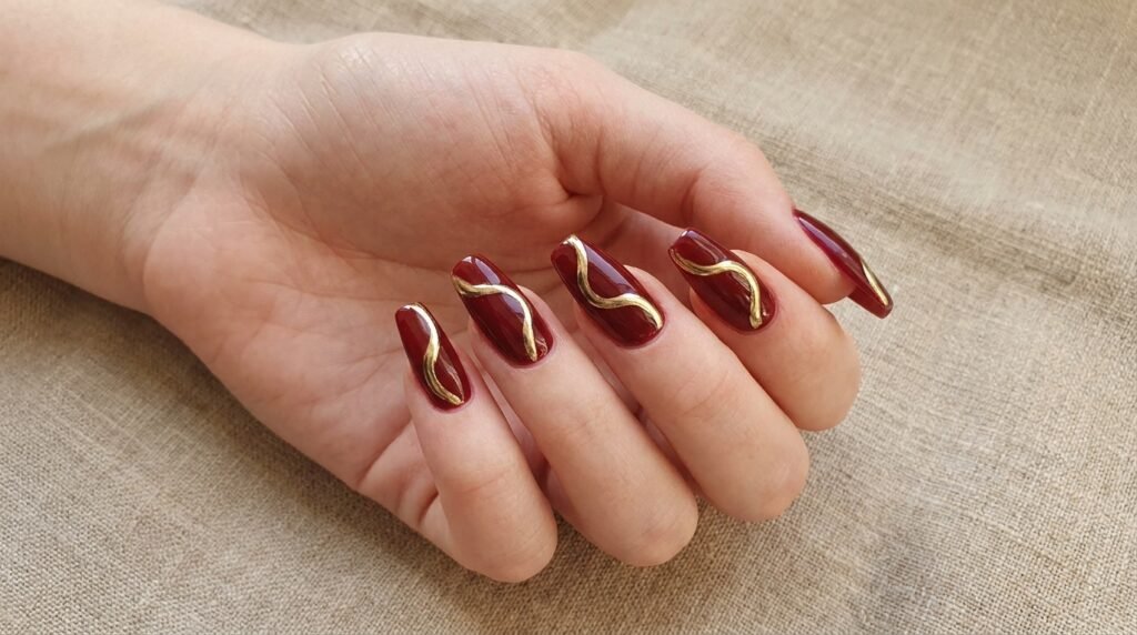 Dark cherry burgundy coffin nails decorated with fluid, abstract metallic gold swirls.