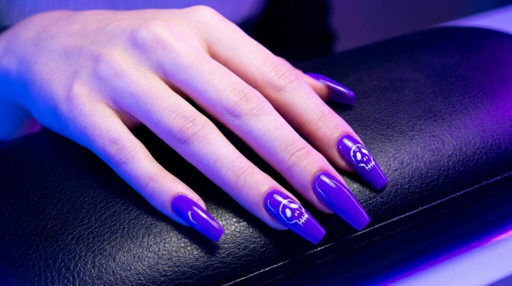 Long coffin nails with white skull artwork over bright neon purple polish on white hand.