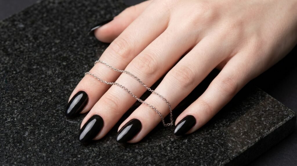 Almond shaped black nails with silver metal chains on hand with very white skin resting on granite.