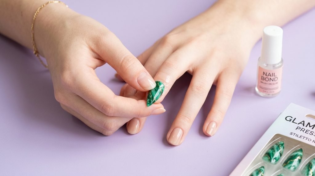 Eighteen year old fair female hand applying an emerald marble press-on stiletto nail over a pastel lavender backdrop.