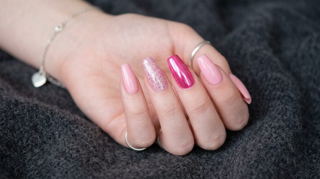 Close-up photograph of an 18-year-old female hand with smooth, very white skin, displaying diverse medium-length coffin nails with plain pink cream, simple glitter, and hot pink chrome finishes, representing press-ons and professional looks, posed against a soft deep charcoal cashmere background.