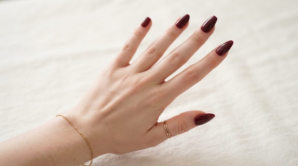A close-up view of five fingers fanned out, each displaying a different nail shape in deep burgundy: squoval, almond, coffin, and stiletto. The background is clean and soft.