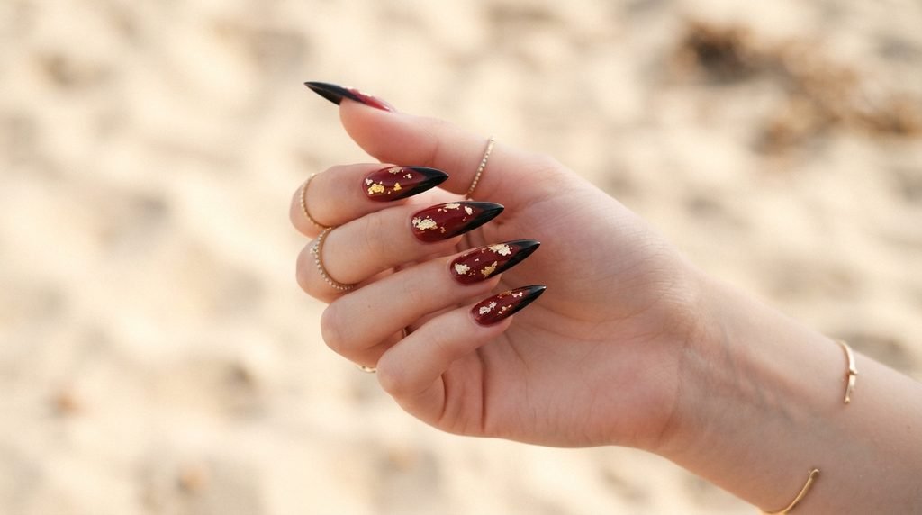 Long oxblood red stiletto nails with black tips and gold foil on an 18-year-old white female hand over sand background.