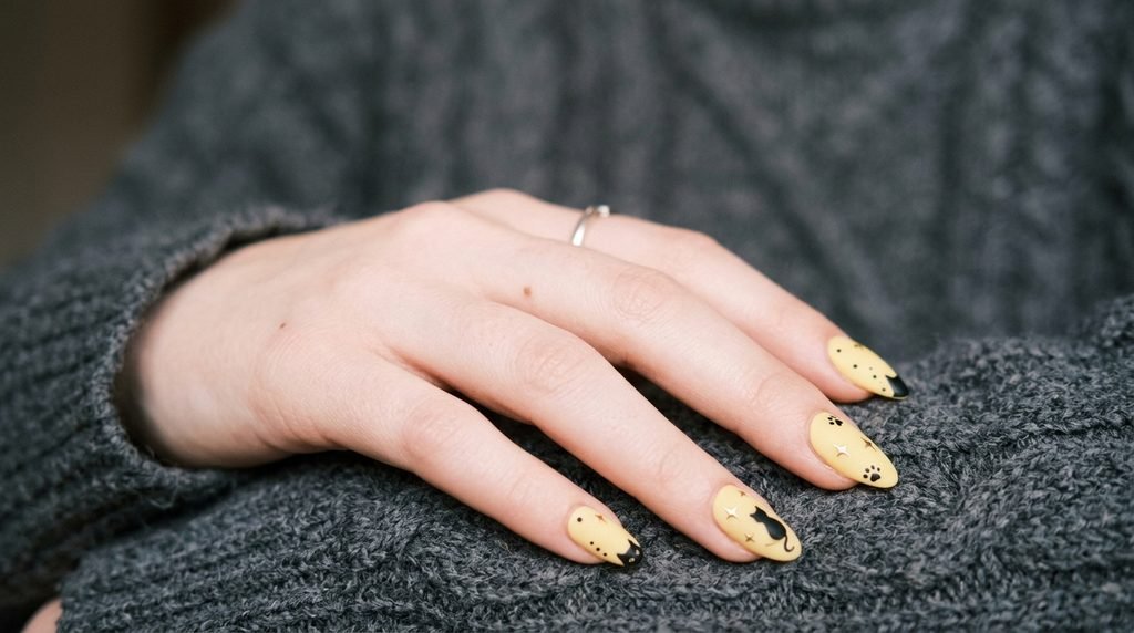 Almond nails with matte butter yellow polish, black cat silhouettes, and tiny star art on white skin.