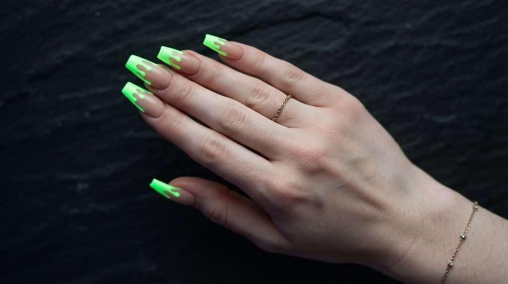 Long coffin nails with nude base and bright glowing neon green dripping French tips on dark slate.
