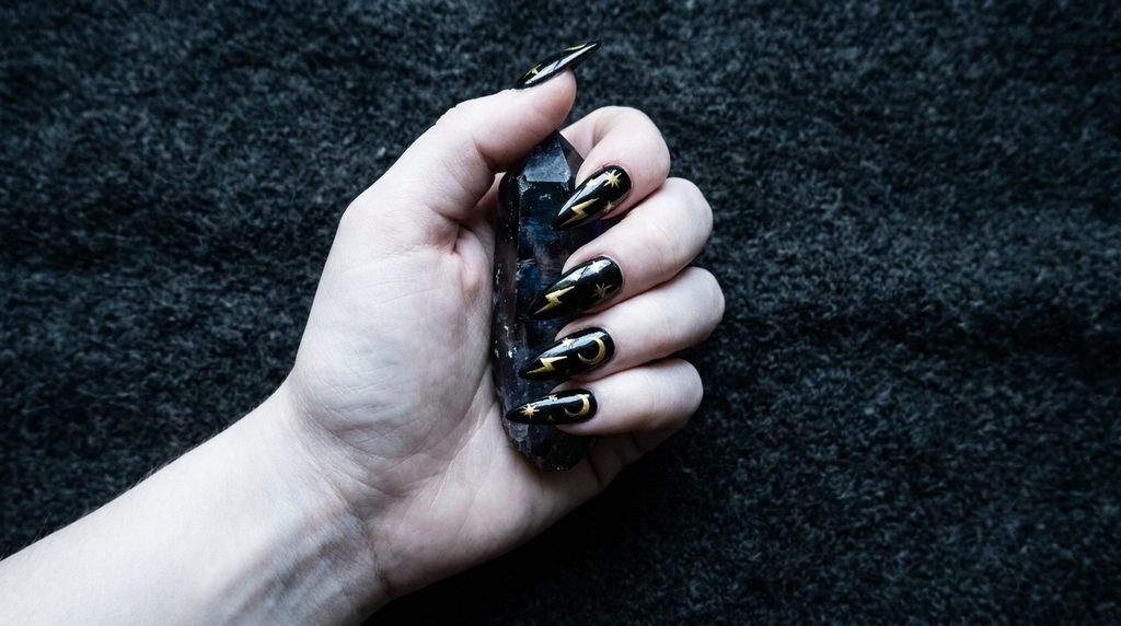 Long stiletto black nails with sharp gold painted lightning bolt and celestial designs flowing towards the point, holding a dark crystal against charcoal background.