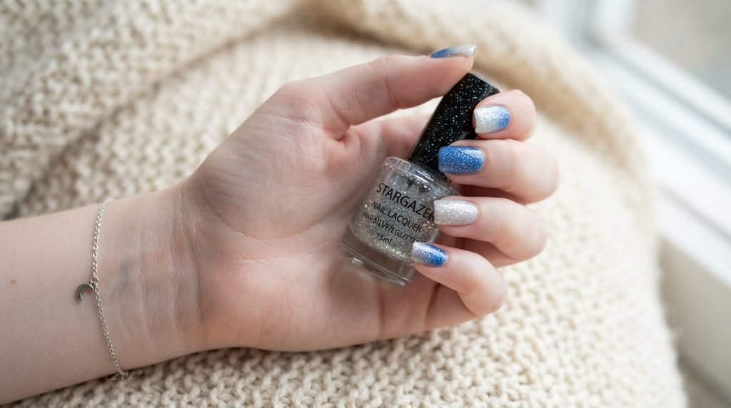 Close-up of a female hand with short square blue glitter ombré nails holding a polish bottle.