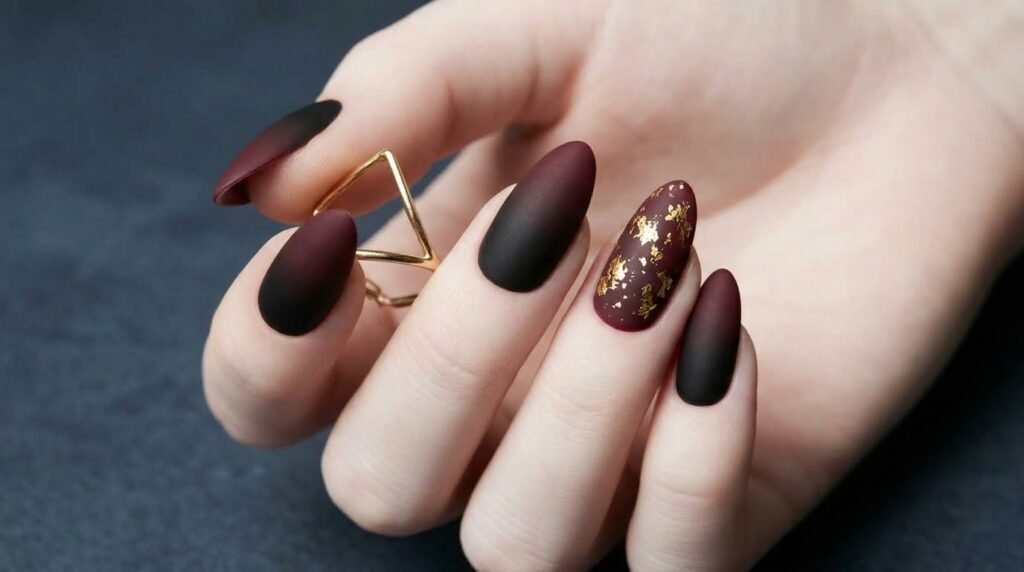 Close-up of matte black and burgundy gradient nails with strategic gold foil flecks, on a white female hand 