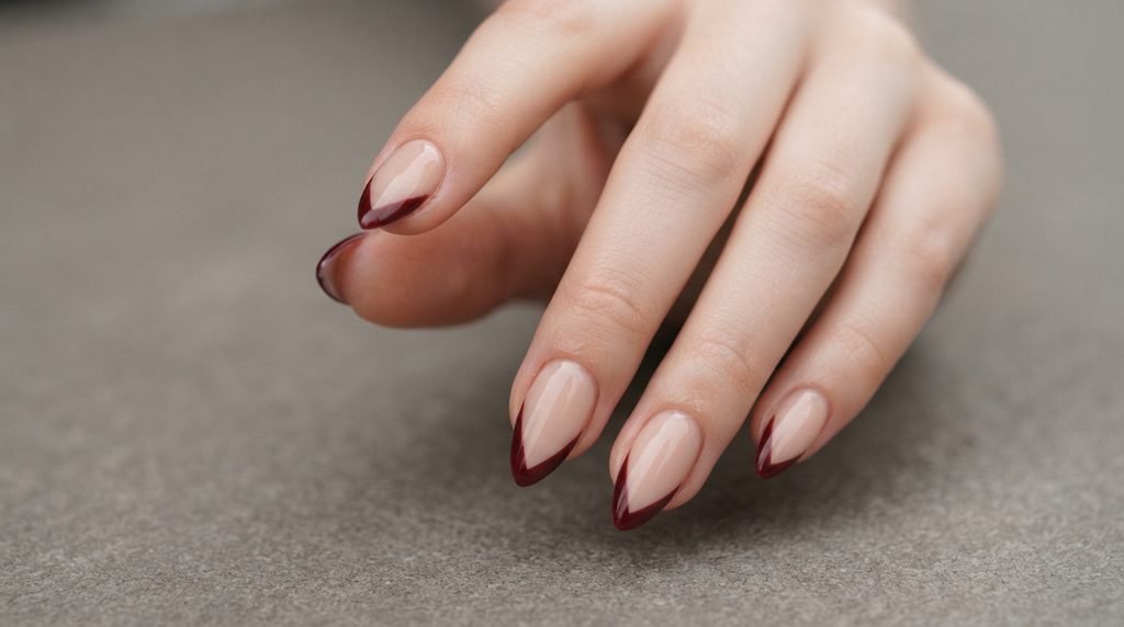 A detailed photograph of medium almond nails with a nude base and sharp V-shaped oxblood French tips on an 18-year-old female hand