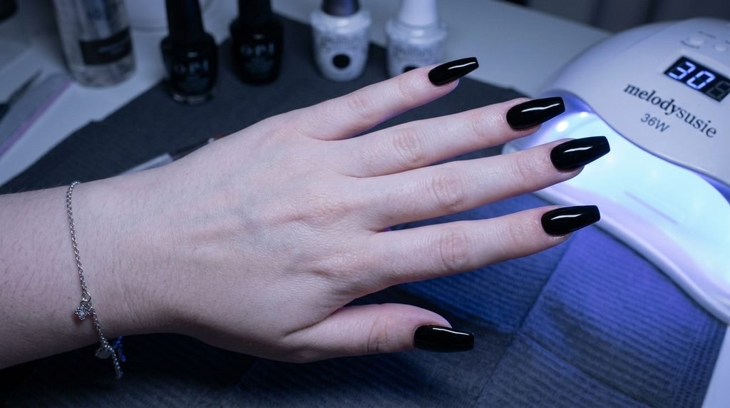 Medium length coffin nails with smooth high-gloss deep black polish, illuminated by the soft blue light from a gel curing lamp nearby.
