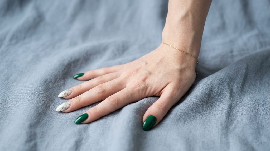 Eighteen year old hand with fair skin showing a balanced mix of solid emerald green and marble accent stiletto nails on slate blue fabric.