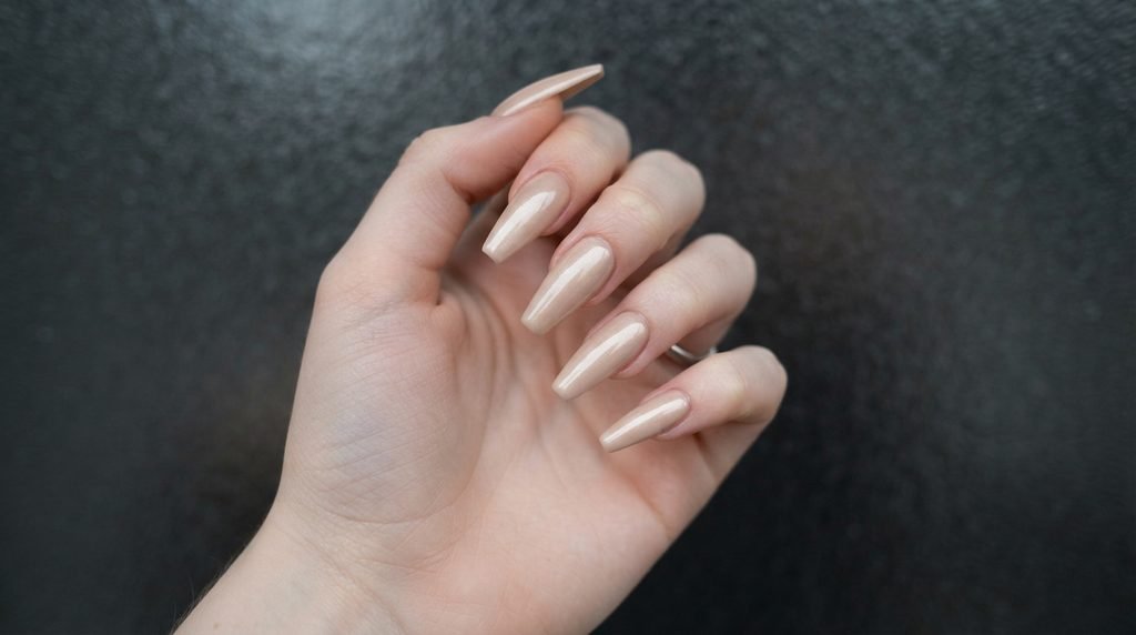 Long champagne beige acrylic coffin nails shown over a dark frosted glass 