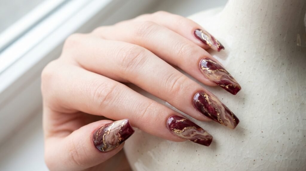 Long coffin nails featuring a detailed burgundy, cream, and gold marble pattern on fair skin.