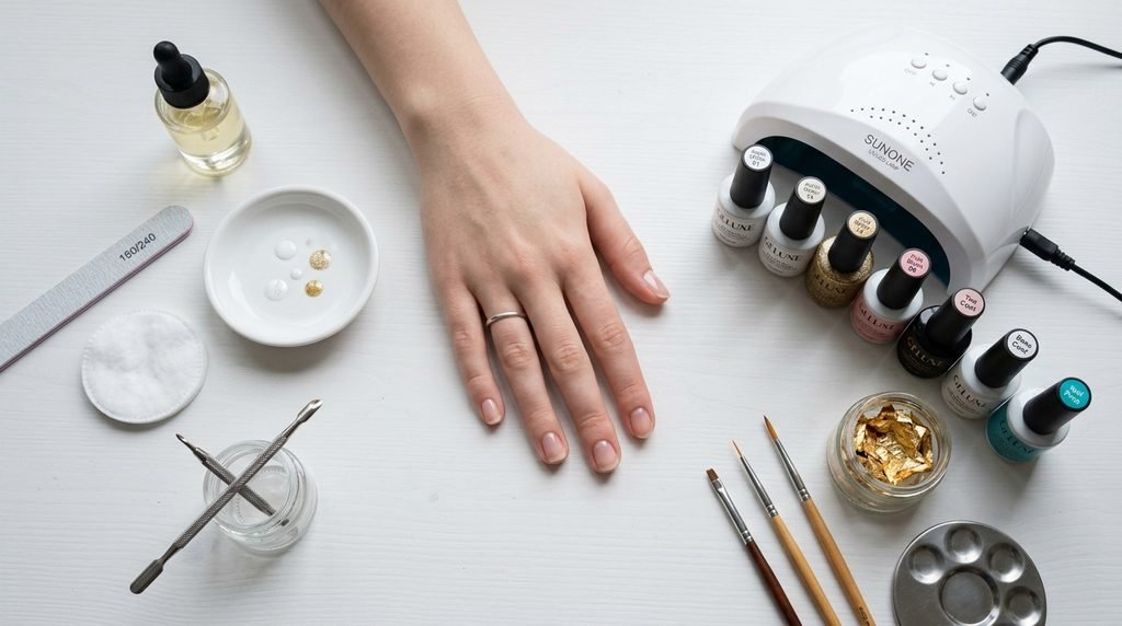 Flat lay of DIY gel nail supplies and an 18-year-old white female hand on a white desk surface.