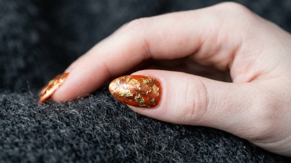 Close-up of gold foil and copper chrome accents on orange nails worn by 18-year-old white hand.