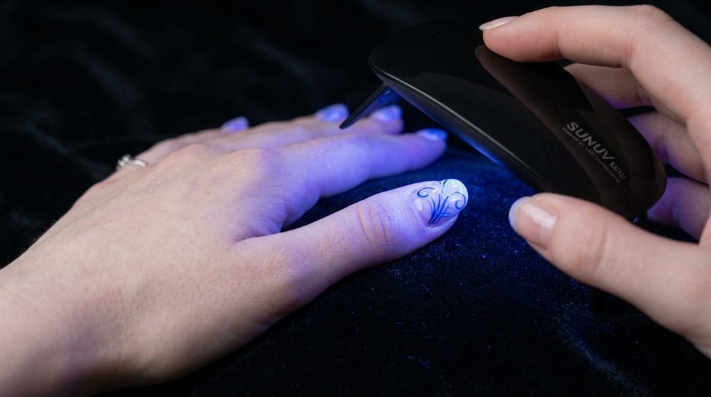 Female hand with white skin flash-curing detailed gel nail art under a mini UV LED lamp over dark velvet.