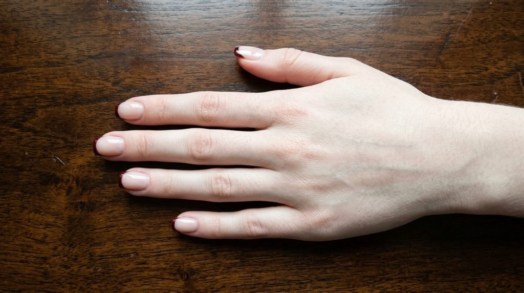 A clean, close-up photograph of short square nails with a very thin oxblood French tip on a nude base."