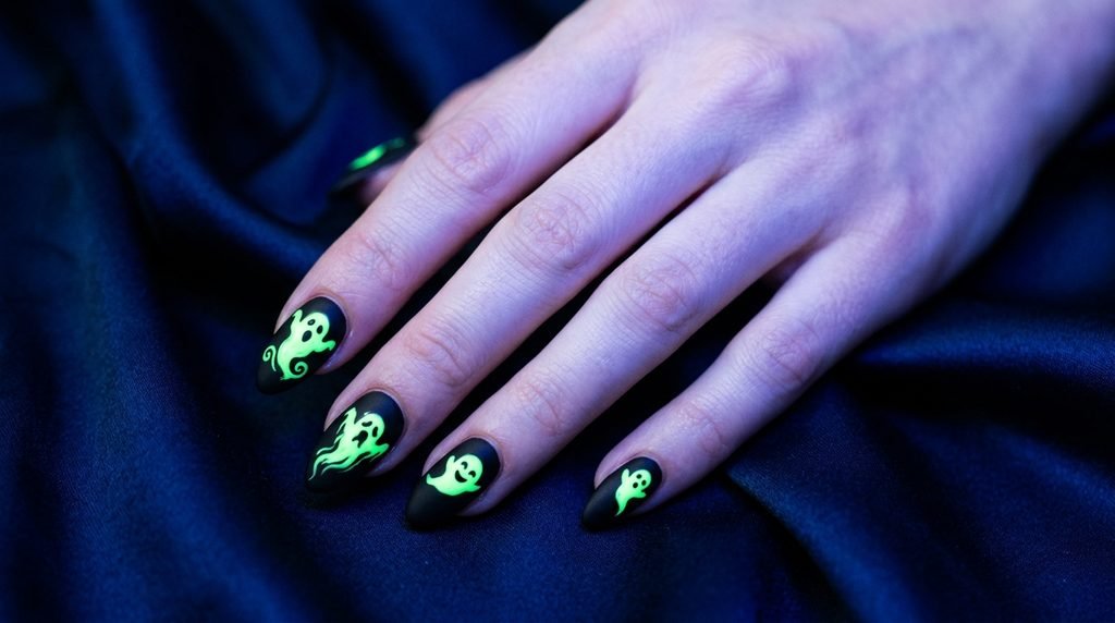 Almond nails painted matte black with bright glowing neon green ghosts on a dark blue silk background.