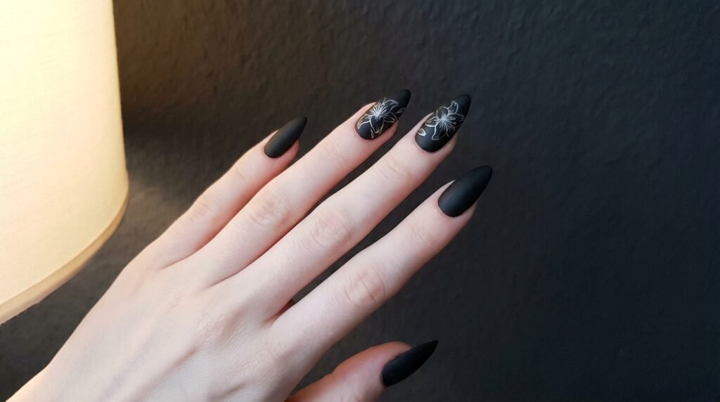 Matte black stiletto nails featuring silver lily art on very white skin.
