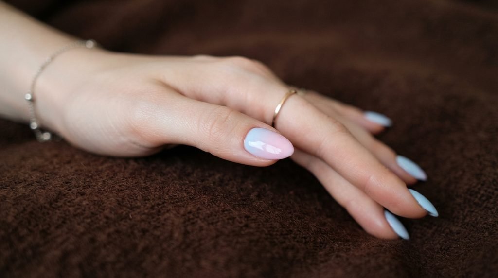 Soft baby blue-to-pink ombre gradient accent nail on a female hand with white skin over chocolate brown.