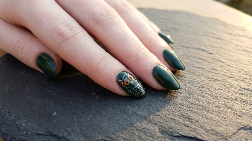 An 18-year-old female hand with very white skin showing a stylized owl illustration on an oval forest green nail.
