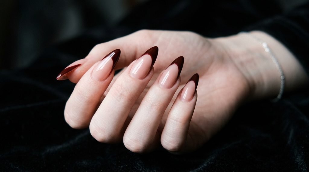 A detailed photograph focusing on a hand with long almond-shaped nails, featuring a steep pointed oxblood French tip