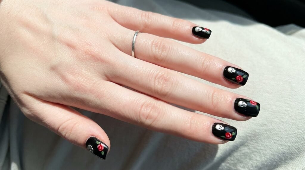 Short square black nails with flat silver skull studs and mini rose art on white skin hand.