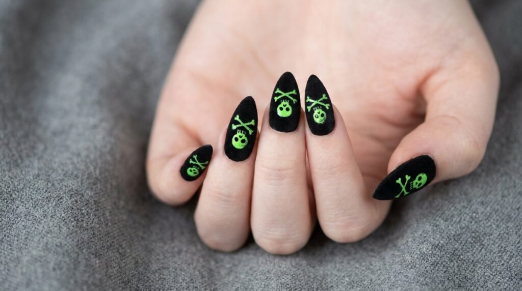 Stiletto nails with green neon skull art over matte black polish on a white skin hand.