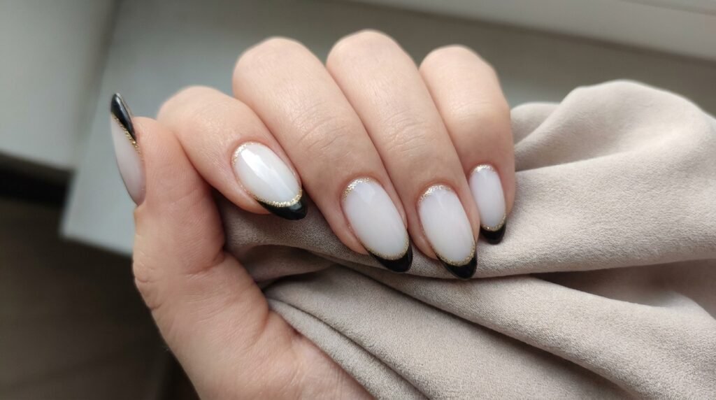 Milky nude almond nails with black French tips outlined by a fine gold glitter line.