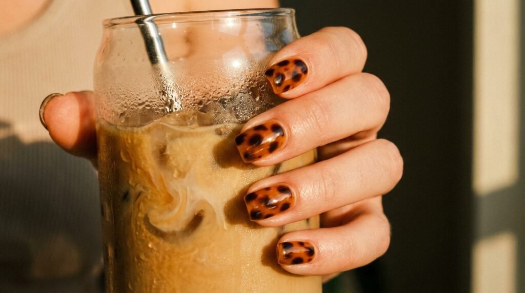 Short squoval tortoiseshell nails holding an iced coffee in warm sunlight.