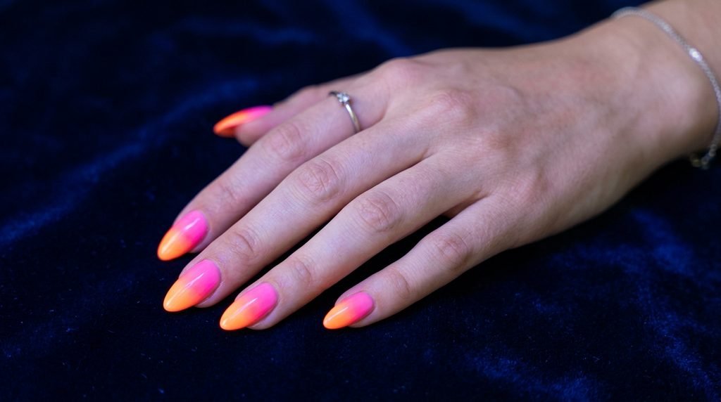 Glowing pink and orange gradient almond nails illuminated against a dark blue background