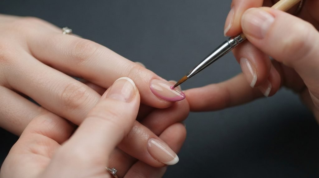 Fine nail brush applying a pink French tip smile line on an almond nail over a charcoal background.