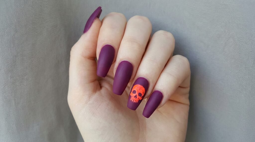 Medium coffin nails with neon orange skull accent over matte plum purple polish on white hand.