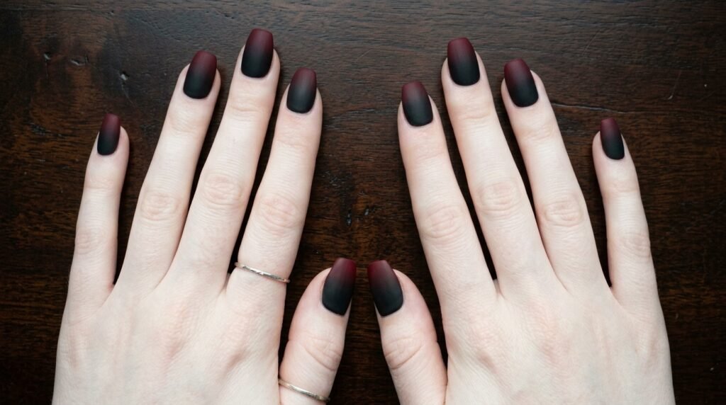 Overhead close-up of reverse burgundy to black matte gradient nails on two white female hands (18), resting on dark wood.