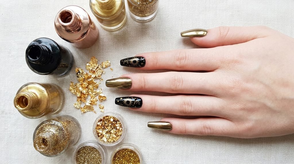 Female hand with medium coffin black and gold tarot nails beside bottles of black and different gold nail polishes and foils.