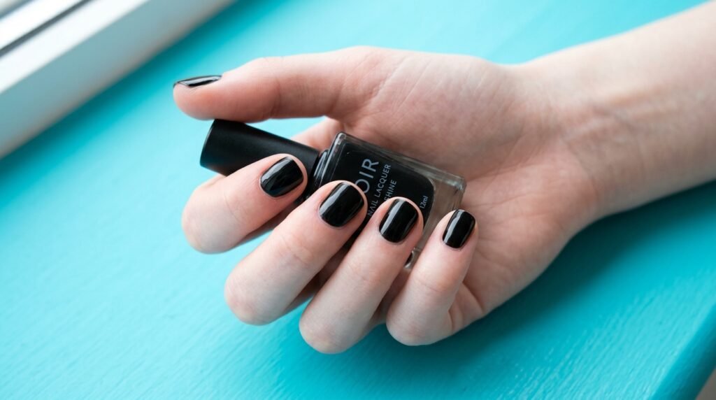 Natural short nails painted in solid glossy black polish on a white skin hand against a turquoise background.