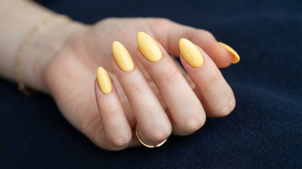 Opaque butter yellow polish on almond nails with white skin tone against dark background.