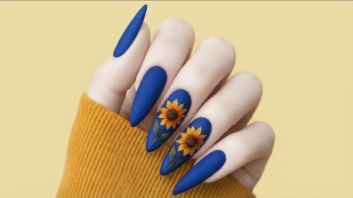 Matte almond nails in deep cobalt blue featuring detailed yellow sunflower nail art on an 18-year-old female hand.