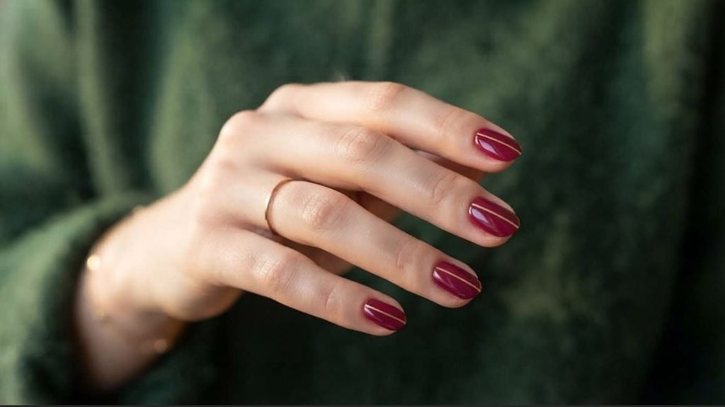A close-up photograph of short almond nails with glossy deep berry base and incredibly thin gold micro vertical lines down the center, against a blurred forest green woolen background.