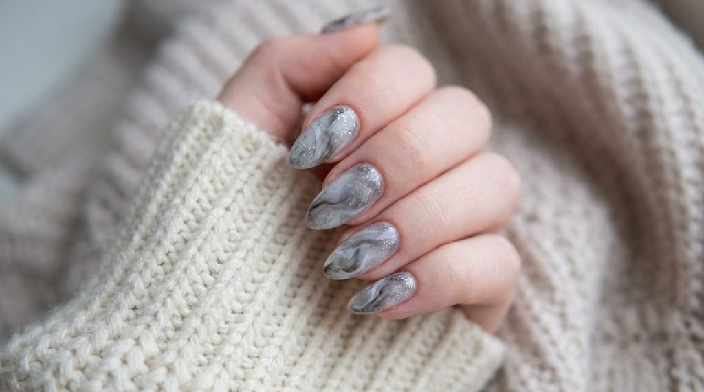 Smoky marble nails with fine silver glitter on an 18-year-old female hand wrapped in a cream sweater.