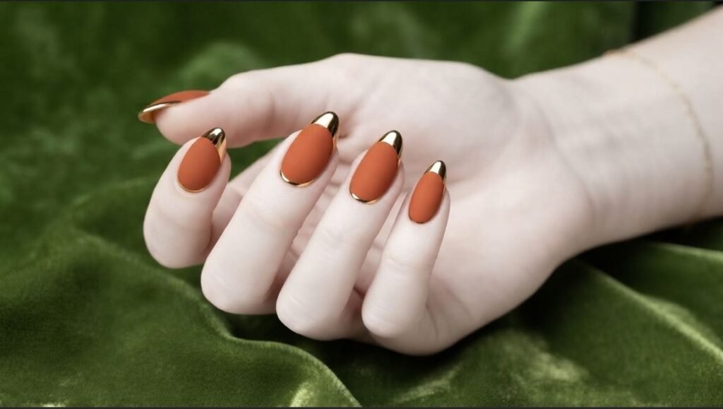 Burnt orange chrome French tips on almond nails, held by 18-year-old female hand with very white skin.