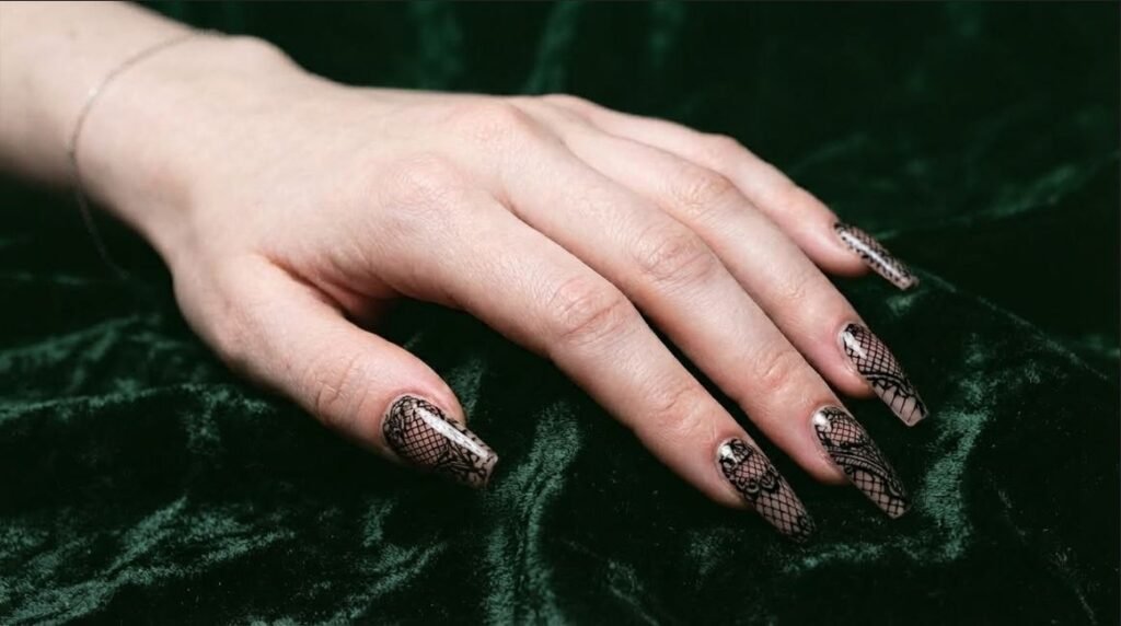Long coffin nails with sheer pink base and detailed black lace nail art on a white skin female hand.