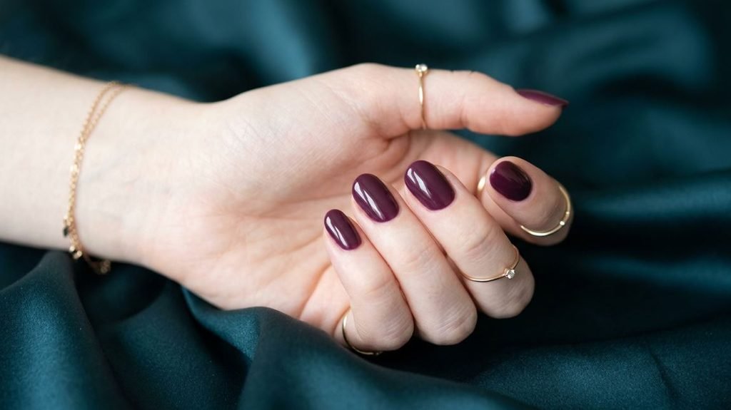 A close-up photograph of short oval nails painted a glossy deep plum color on an 18-year-old female hand with very white skin, against a blurred dark teal silk background.