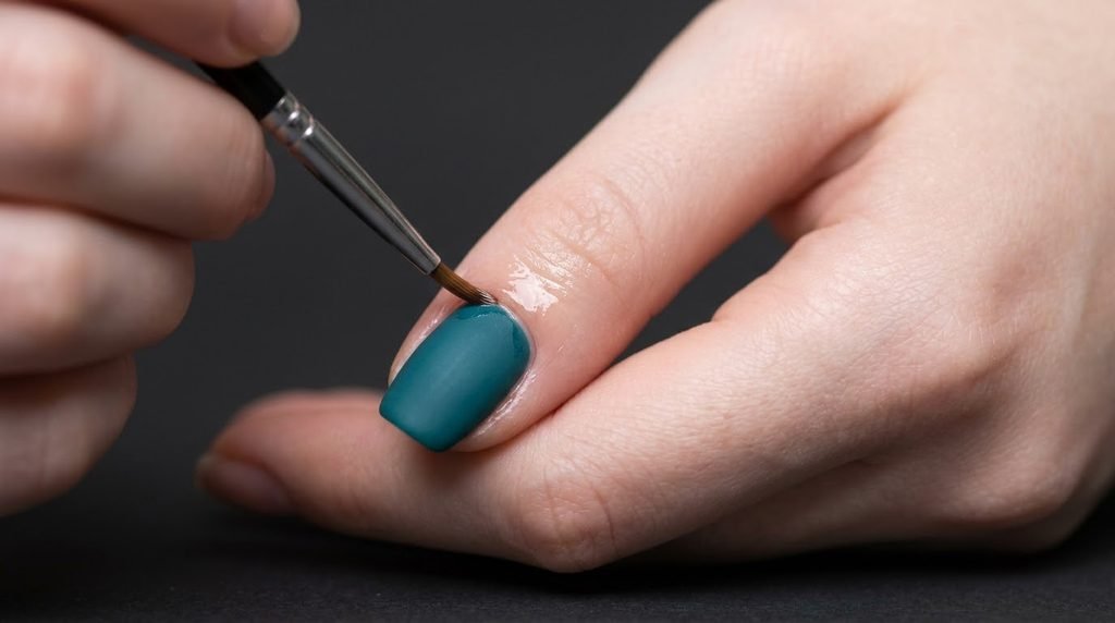 An 18-year-old female hand with very white skin applying cuticle oil precisely around the skin of a matte teal nail against a dark backdrop.