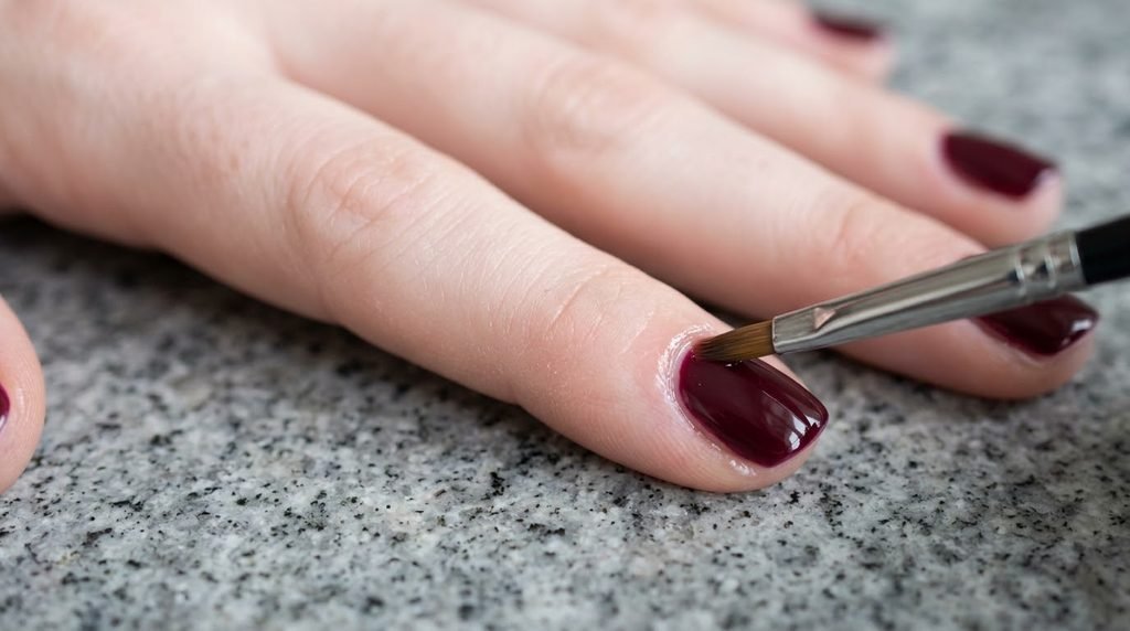 Macro photograph of a detail brush cleaning up dark red polish around a clean cuticle line on fair skin
