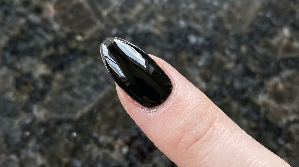 Macro image of a clear top coat being applied over the edge of a glossy black nail on a pale hand.