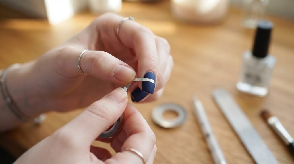 A close-up of a hand with very white skin and short square matte navy nails. One nail has metallic striping tape applied, illustrating a DIY geometric pattern technique.