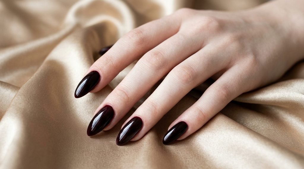 Long elegant almond nails coated in glossy black cherry gel posed on champagne gold satin.